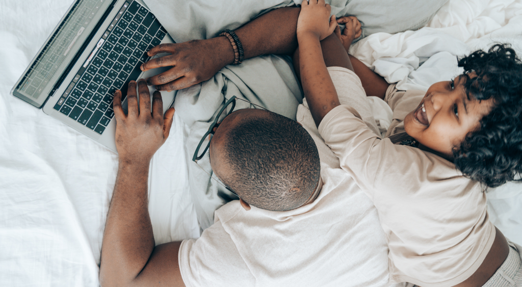 man working on laptop in bed with child climbing on his back